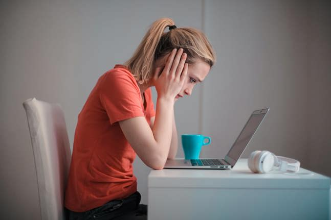 Blonde haired woman sitting at a desk wearing a red top while looking at a laptop with her head in her hands.