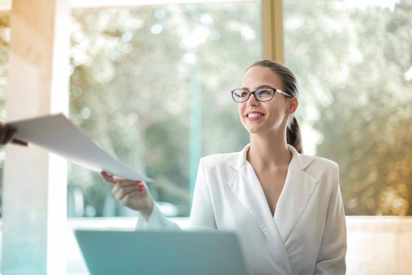 A woman wearing a white shirt and glasses sitting at a desk handing a piece of paper over to another person