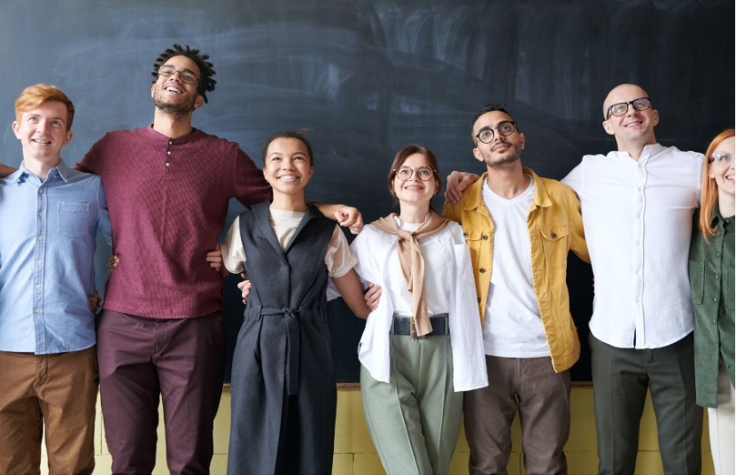 A group of 7 people standing in a line smiling with their arms around each other